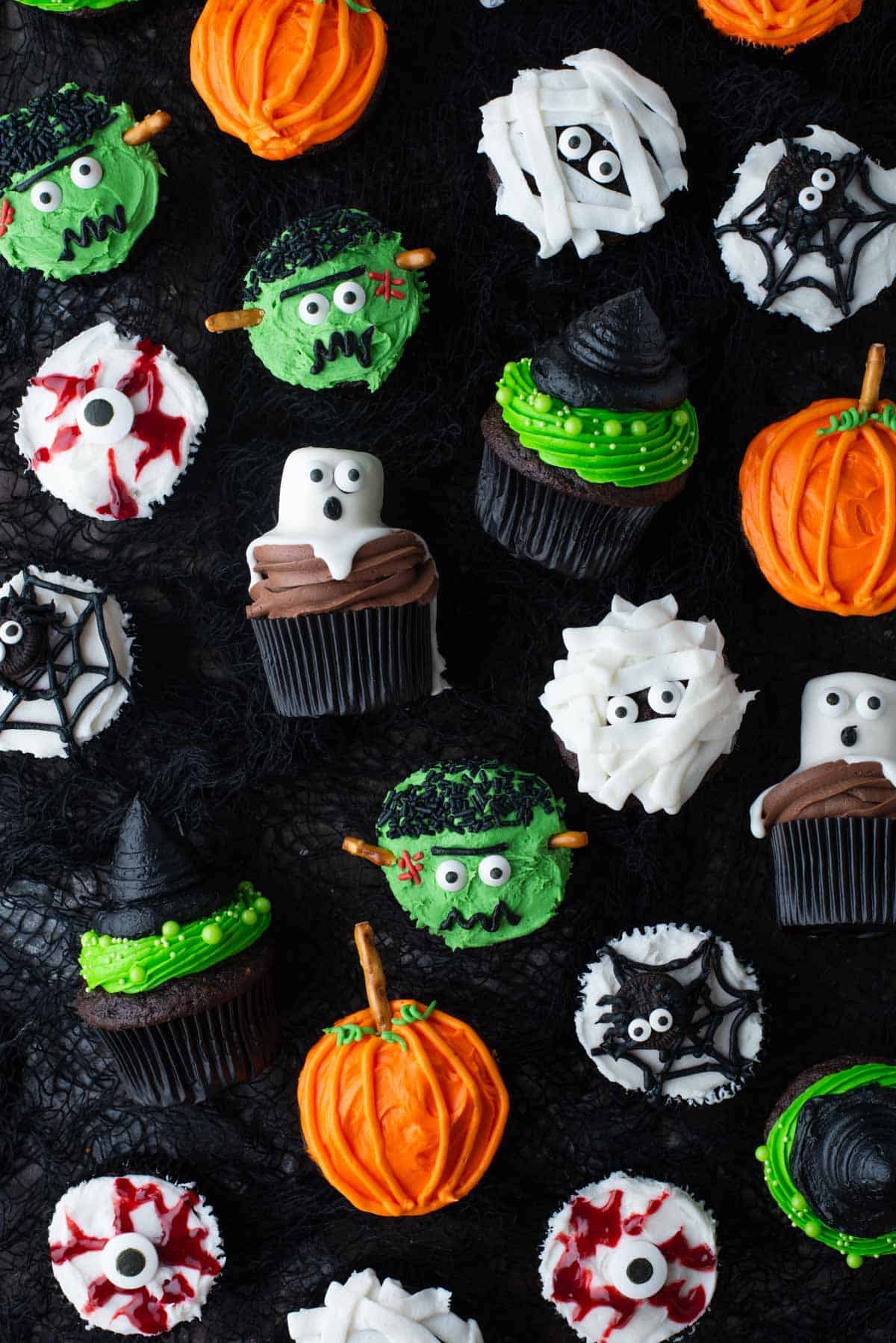 an assortment of halloween cupcakes including mummy cupcakes, pumpkin cupcakes, spider web cupcakes, witch hat cupcakes and ghost cupcakes