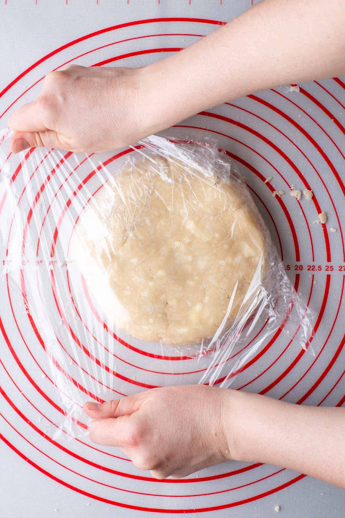 a disc of pie crust dough sitting on a red and white pie mat being wrapped with clear plastic wrap