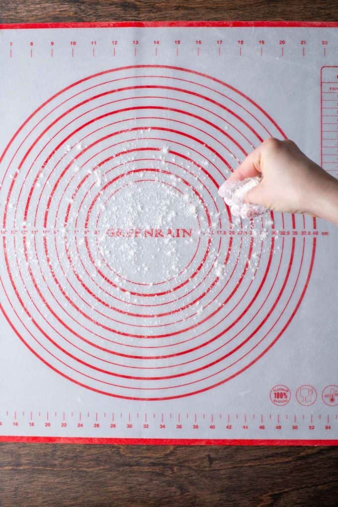 a hand dusting a red and white pie mat that is sitting on a wood surface with flour