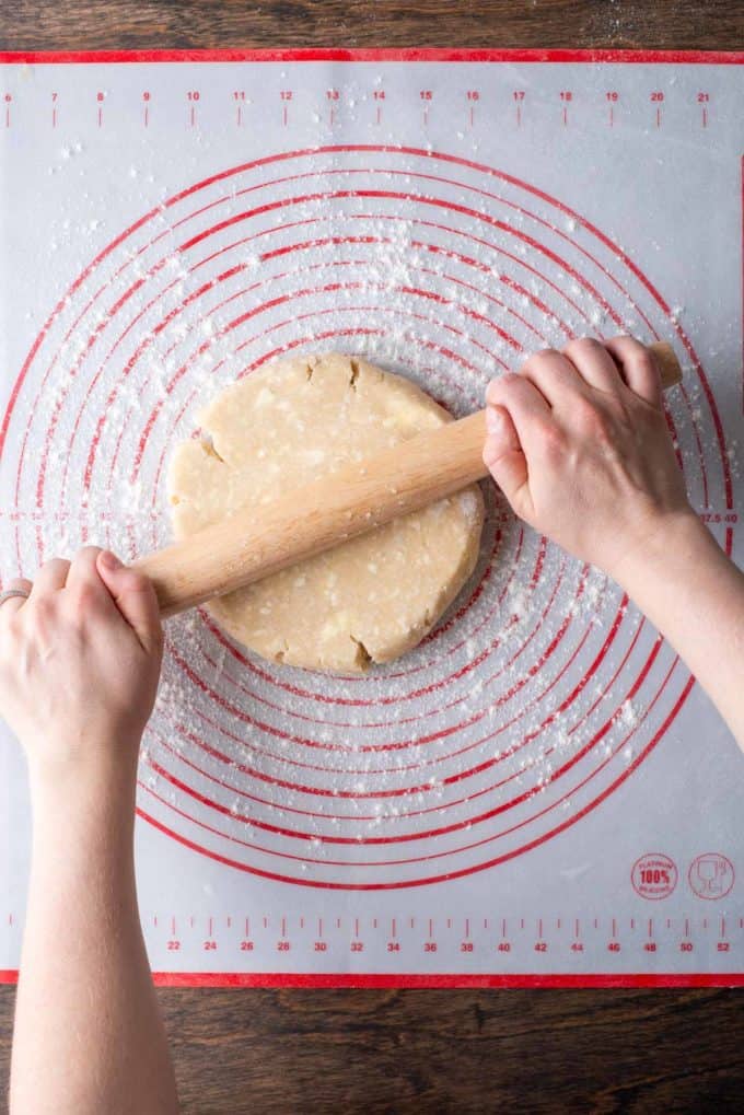 a disc of pie crust dough on a red and white pie mat being rolled with a rolling pin and flour sprinkled beneath it