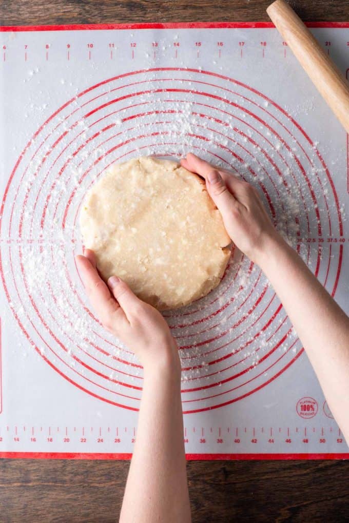 a disc of pie crust dough being placed on a red and white pie mat that is sitting on a wood surface and has been sprinkled with flour, with a wooden rolling pin to the side