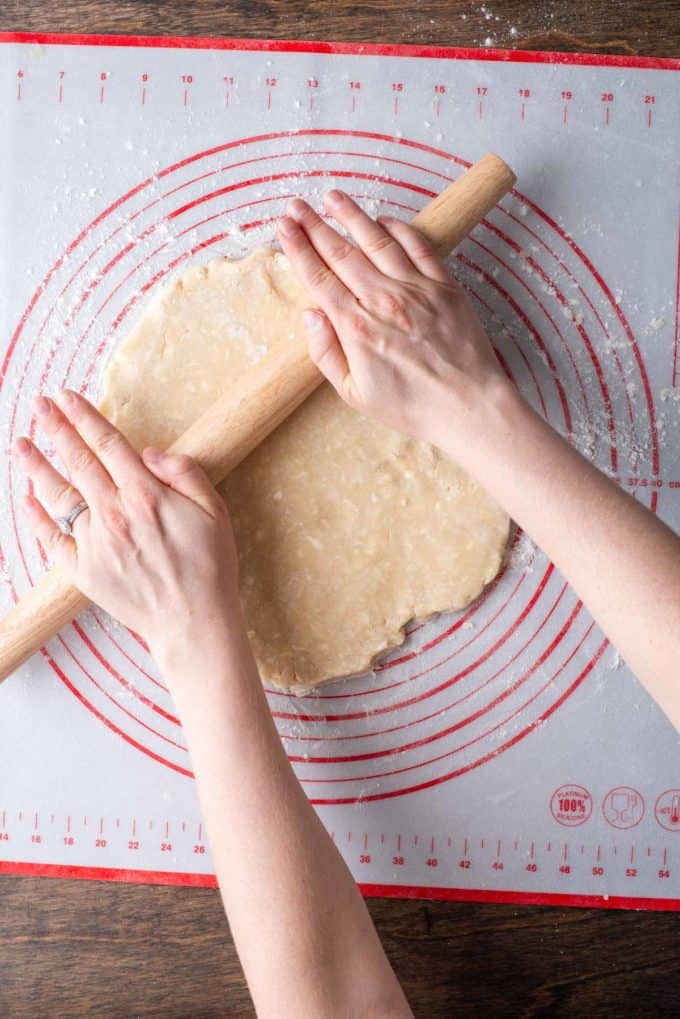 a disc of pie crust dough on a red and white pie mat being rolled with a rolling pin and flour sprinkled beneath it