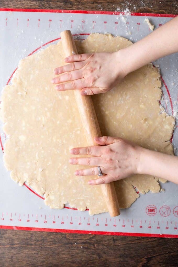 a large round imperfect circle of pie crust dough being rolled out with a wood rolling pin on top of a red and white pie mat that is sitting on a wood surface and sprinkled with flour