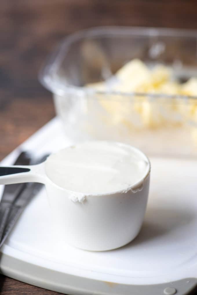 shortening in a white measuring cup on a white cutting board with two butter knives and a dish of butter cubes