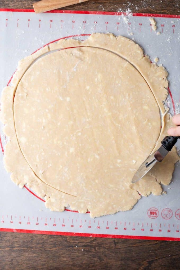 a large circle of pie crust dough being cut with a pizza cutter on top of a red and white pie mat that is sitting on a wood surface and sprinkled with flour