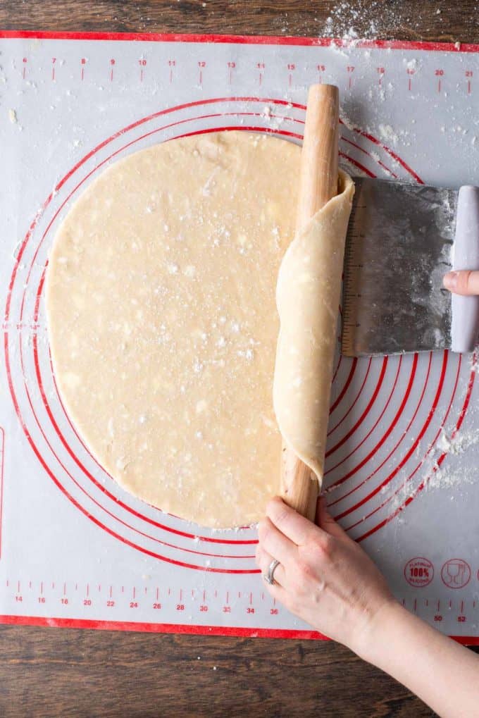 pie crust dough that has been rolled out being lifted by a pastry cutter and rolled onto a wood rolling pin off of a red and white pie mat