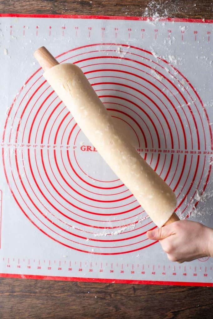 pie crust dough that has been rolled out, trimmed and rolled onto a wood rolling pin held over a red and white pie mat sprinkled with flour