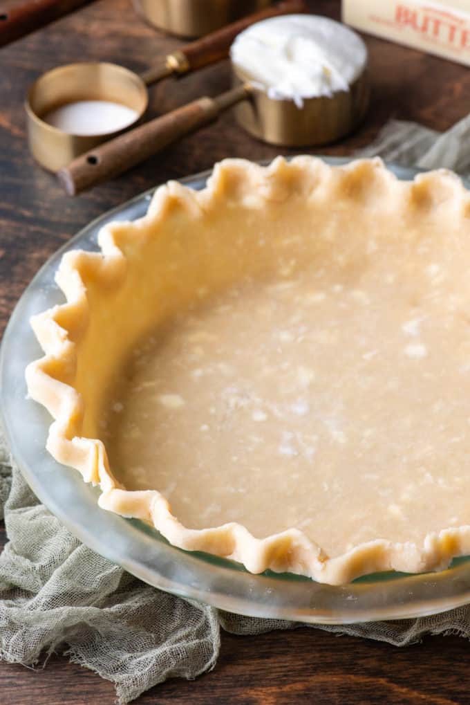 pie crust in a glass pie dish on a wooden surface with a light green towel and ingredients in the background in measuring cups