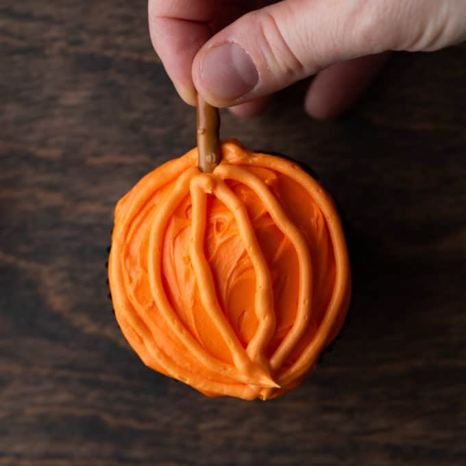 a pumpkin patch cupcake with orange frosting on a wood surface with a pretzel stick being stuck in the top as the stem on the pumpkin