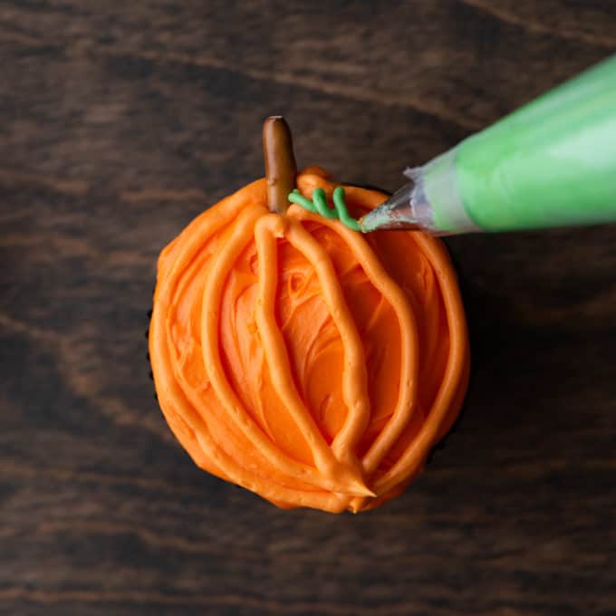 a pumpkin patch cupcake with orange frosting on a wood surface with a pretzel stick sticking out of the top being piped with little green vines beside the pretzel
