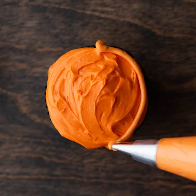 a cupcake with orange frosting on it on a wood surface being piped with an orange line along the right side