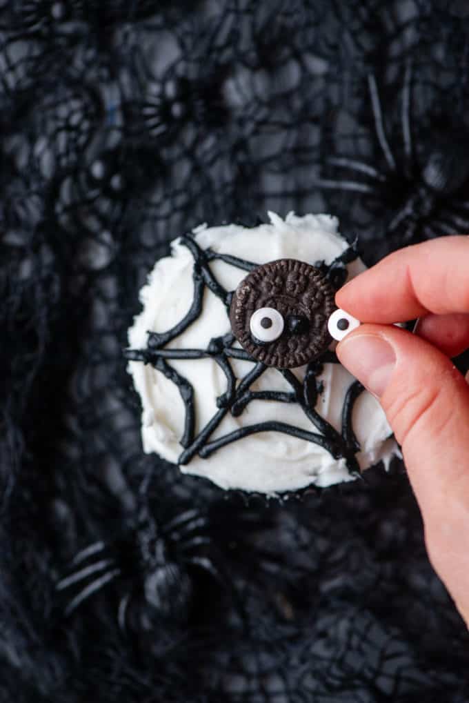 adding mini candy eyeballs on top of a mini oreo to create a spider on top of spider web cupcakes