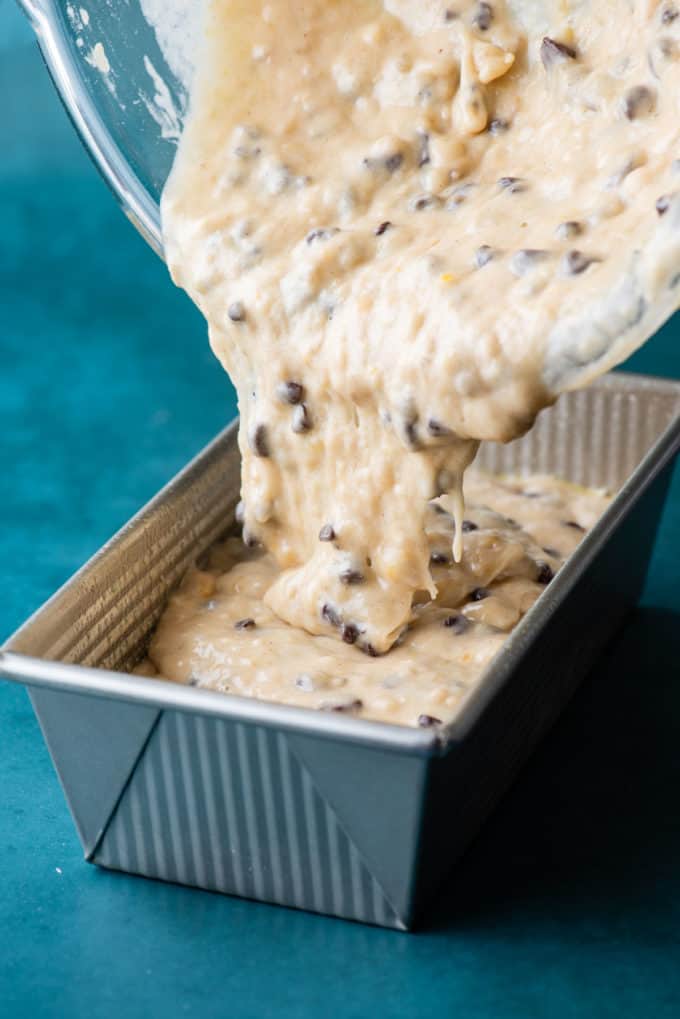chocolate chip banana bread batter being poured into a loaf pan on a dark teal surface
