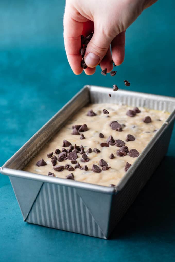 Chocolate Chip Banana Bread batter in a silver loaf pan being sprinkled with more chocolate chips, sitting on a dark teal surface
