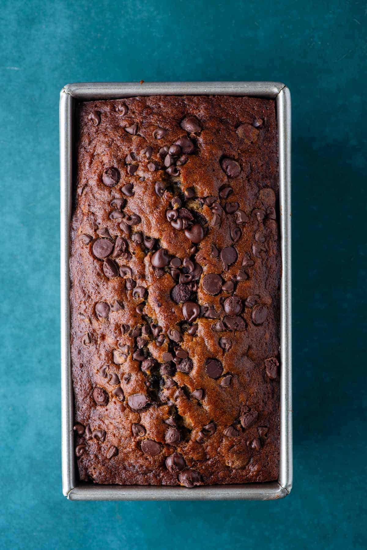 chocolate chip banana bread baking in a loaf pan sitting on a dark teal surface