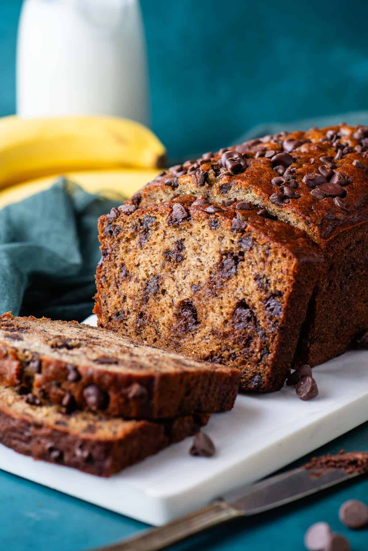 a loaf of chocolate chip banana bread on a white cutting board with a dark teal towel beside is, chocolate chips sprinkled around, and whole bananas and a glass of milk in the background