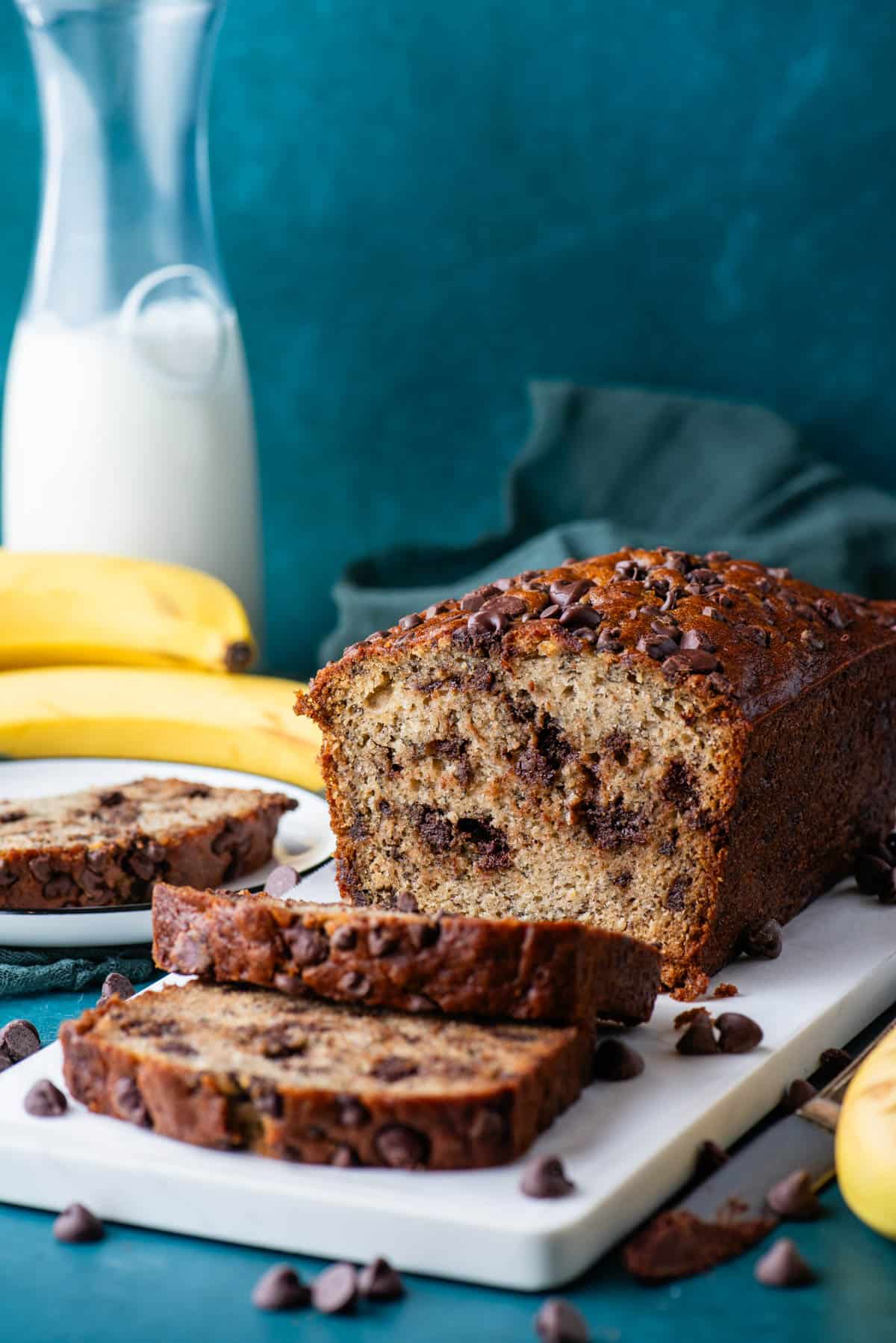 a loaf of chocolate chip banana bread on a white cutting board with a plate with one slice of banana bread beside it, chocolate chips sprinkled around, and whole bananas and a glass of milk in the background