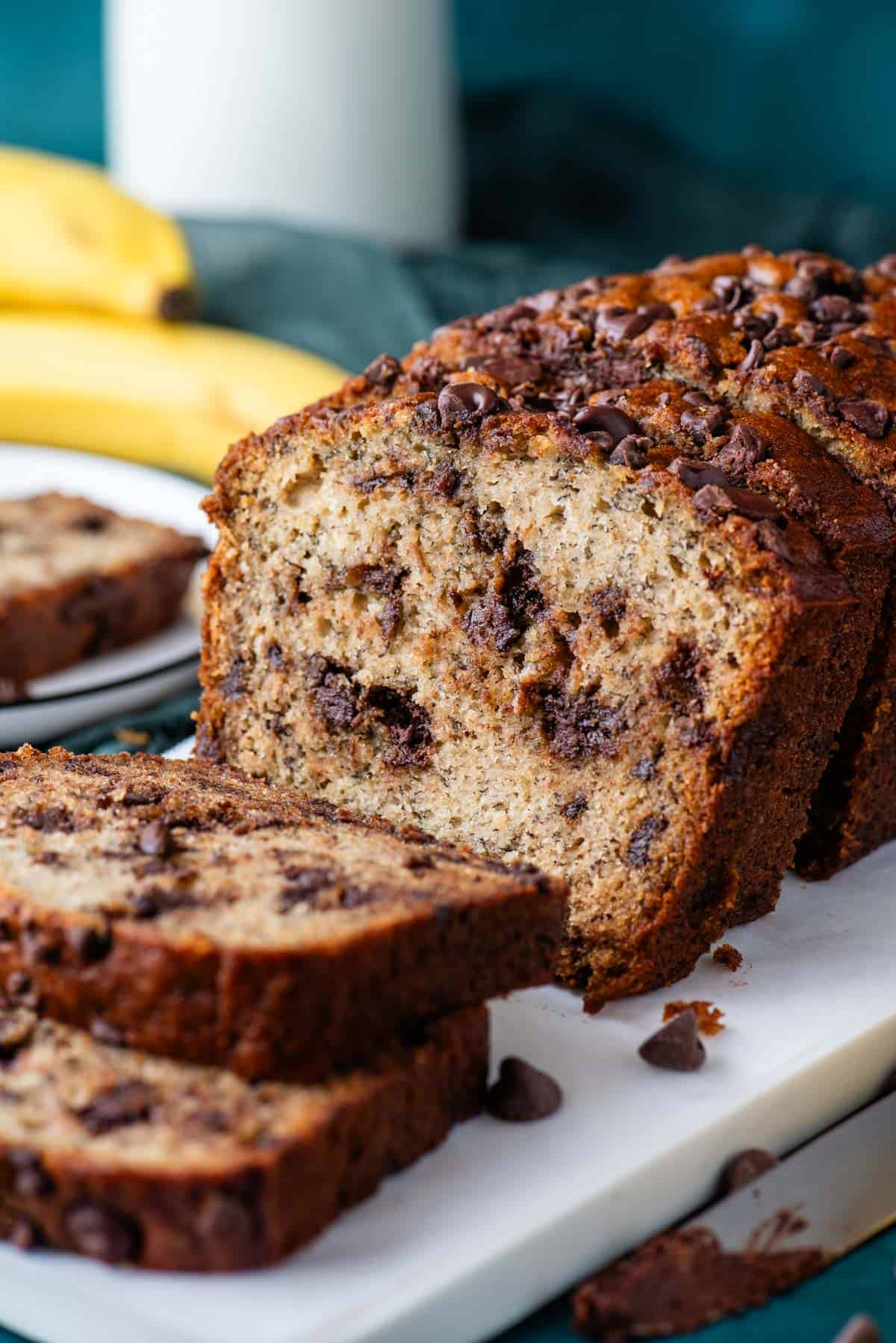 sliced chocolate chip banana bread on a white cutting board with a plate in the background with one slice on it and whole bananas behind that