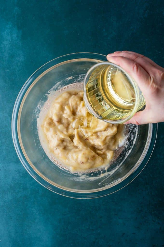 mashed bananas in a clear glass bowl on a dark teal surface with a glass dish of vegetable oil being poured in