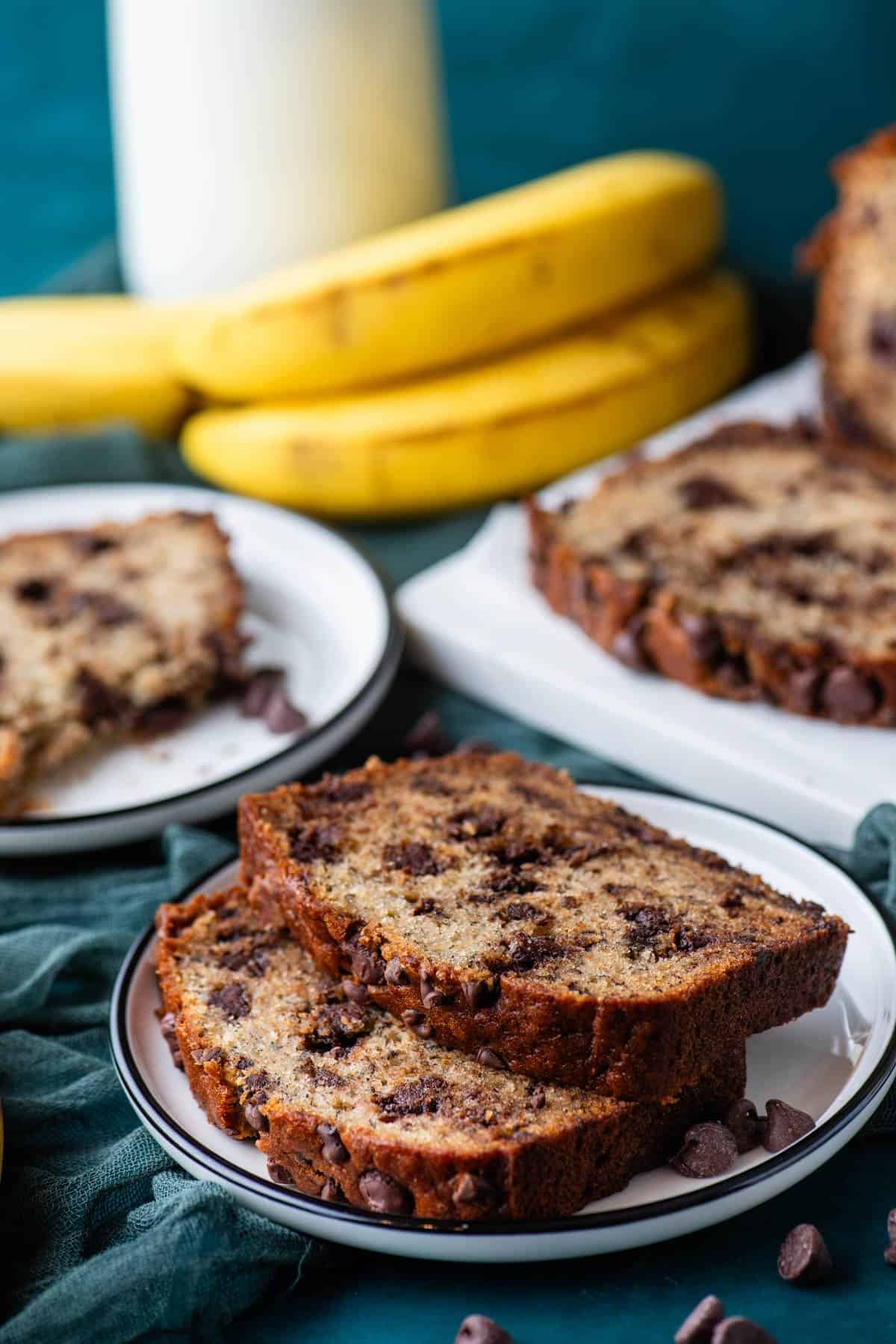 two slices of chocolate chip banana bread on a white plate on top of a dark teal towel, with whole bananas beside it, another plate with a slice of banana bread, and a white cutting board with more bread on it behind it