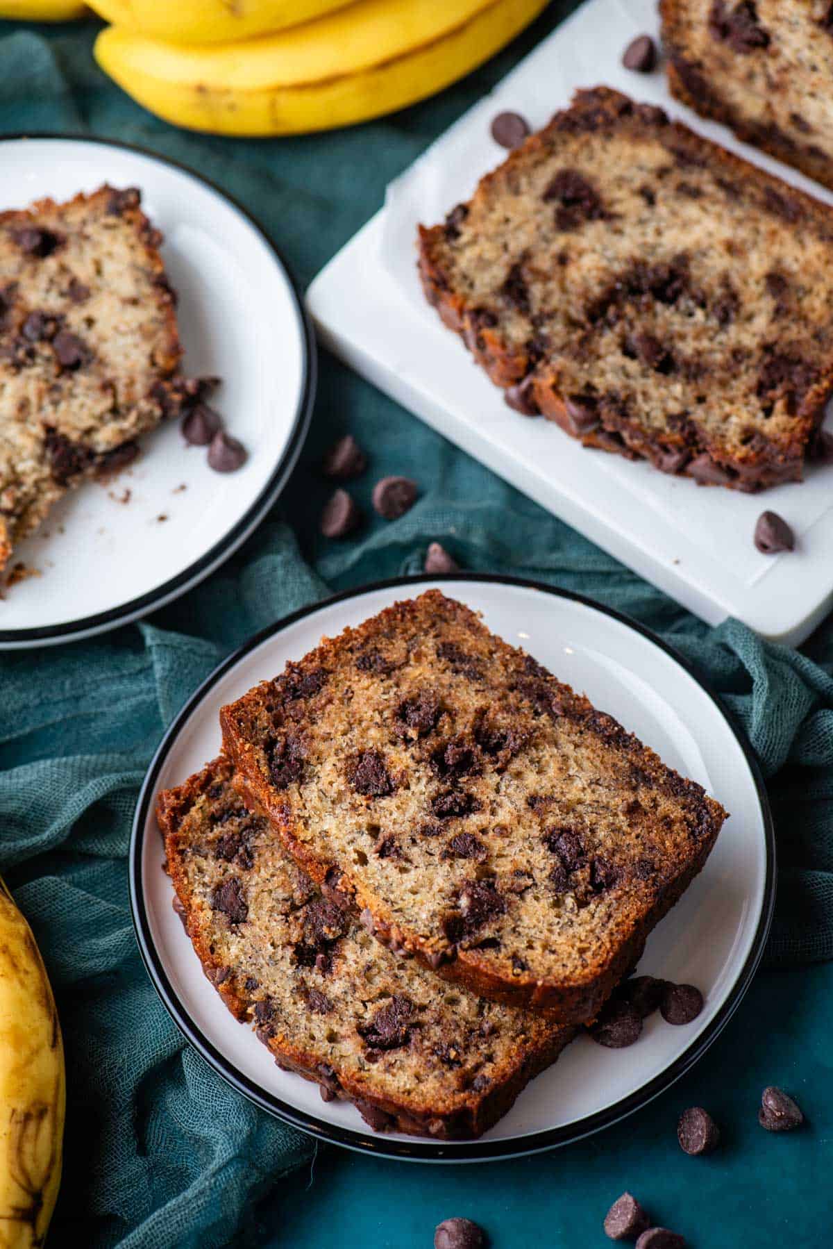 two small white plates with banana bread slices on them, the front one with two slices on it, and a white cutting board with banana bread slices on it, all sitting on a dark teal towel with chocolate chips scattered around