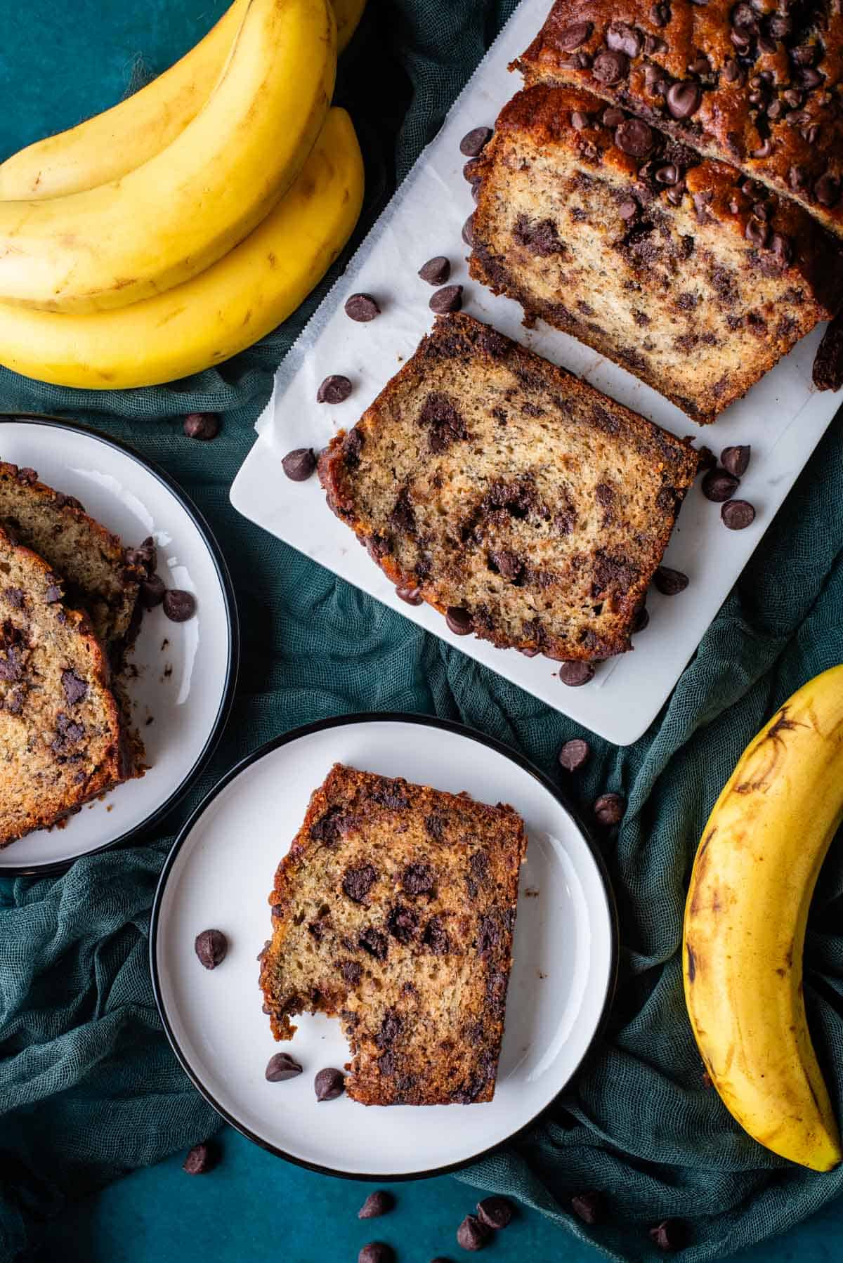 an overhead view of two white plates and one white cutting board with slices of chocolate chip banana bread on them, all sitting on top of a dark teal towel with chocolate chips and whole bananas scattered around