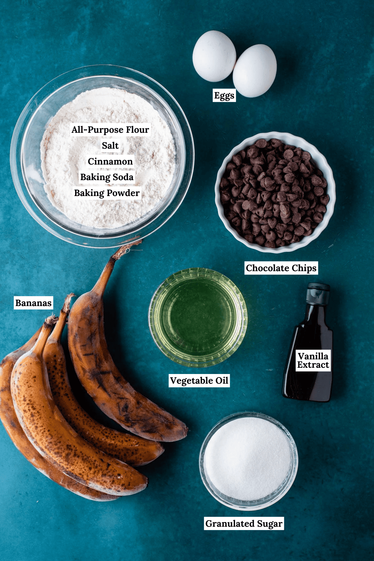 ingredients for chocolate chip banana bread including flour, salt, cinnamon, baking powder, baking soda, eggs, chocolate chips, vegetable oil, vanilla extract, overripe bananas, and granulated sugar