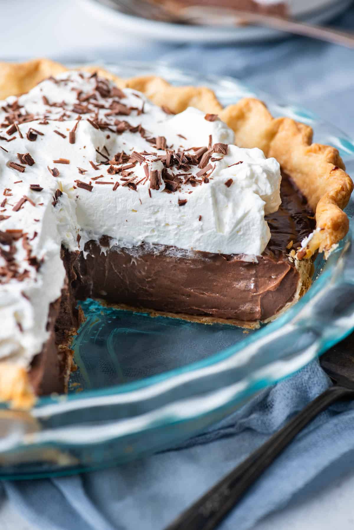 a chocolate pie in a glass pie dish missing one slice of pie, sitting on top of a blue towel with a serving utensil beside the pie dish
