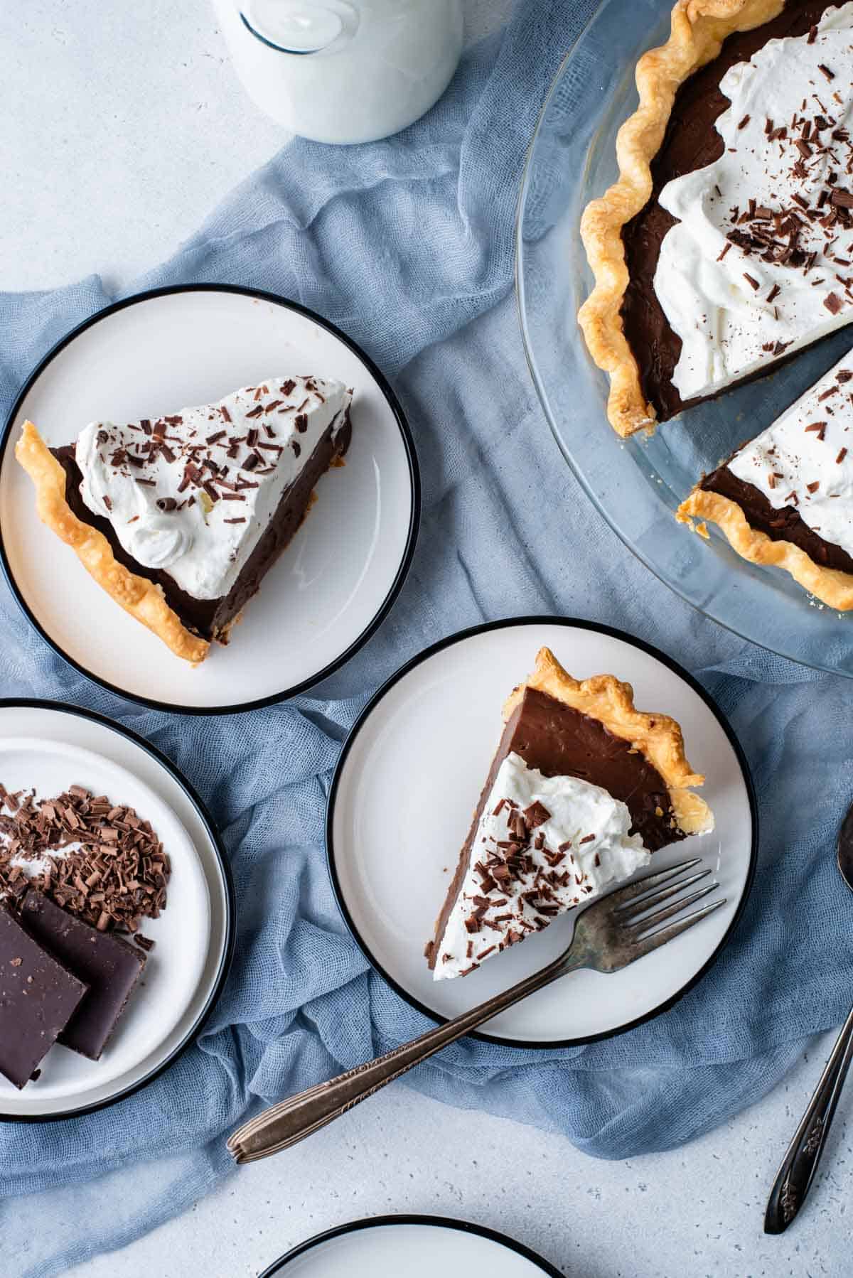 overheard view of a spread of chocolate pie with a glass pie dish with pie slices in it, two small white plates with pie slices on them and a white bowl of chocolate shavings, all sitting on a blue towel