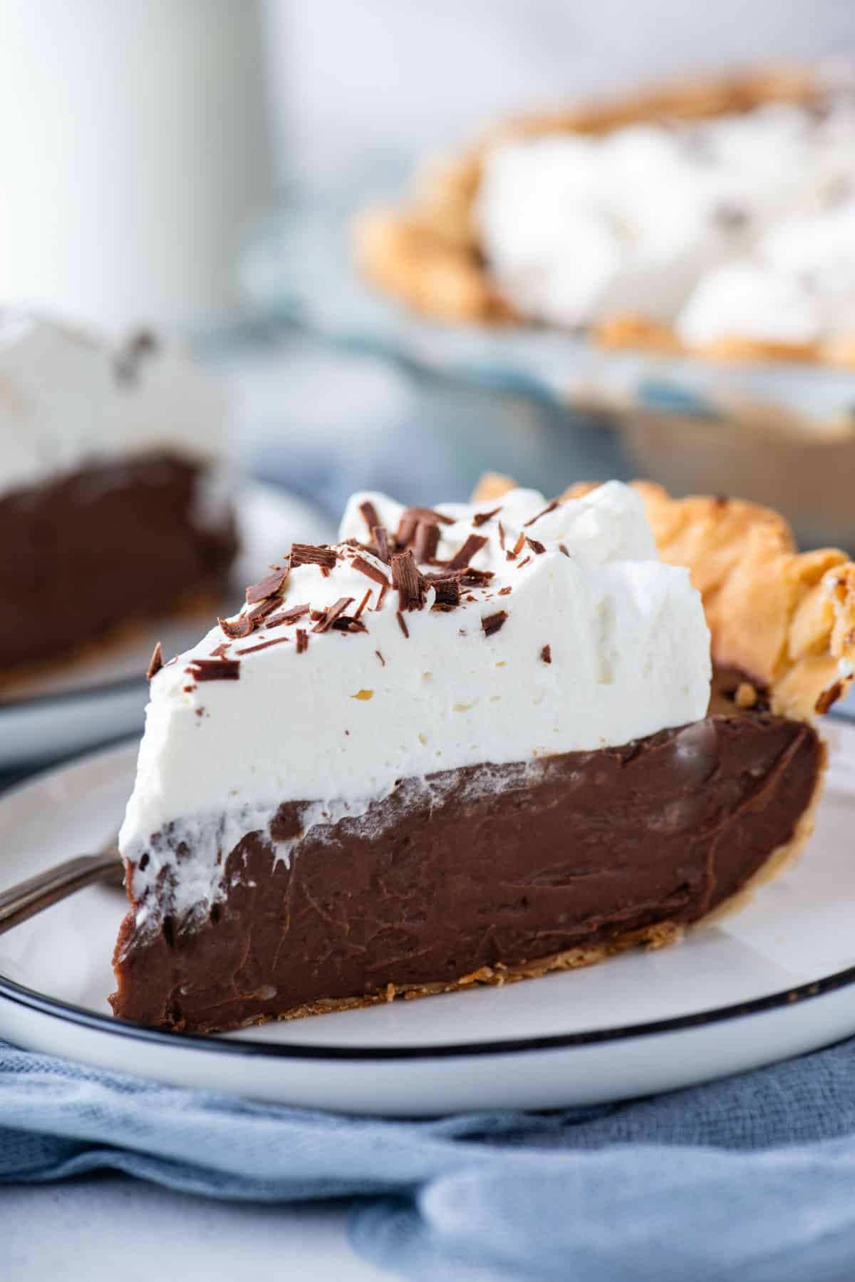 the side view of a slice of chocolate pie on a white plate sitting on a blue towel and more pie in the background