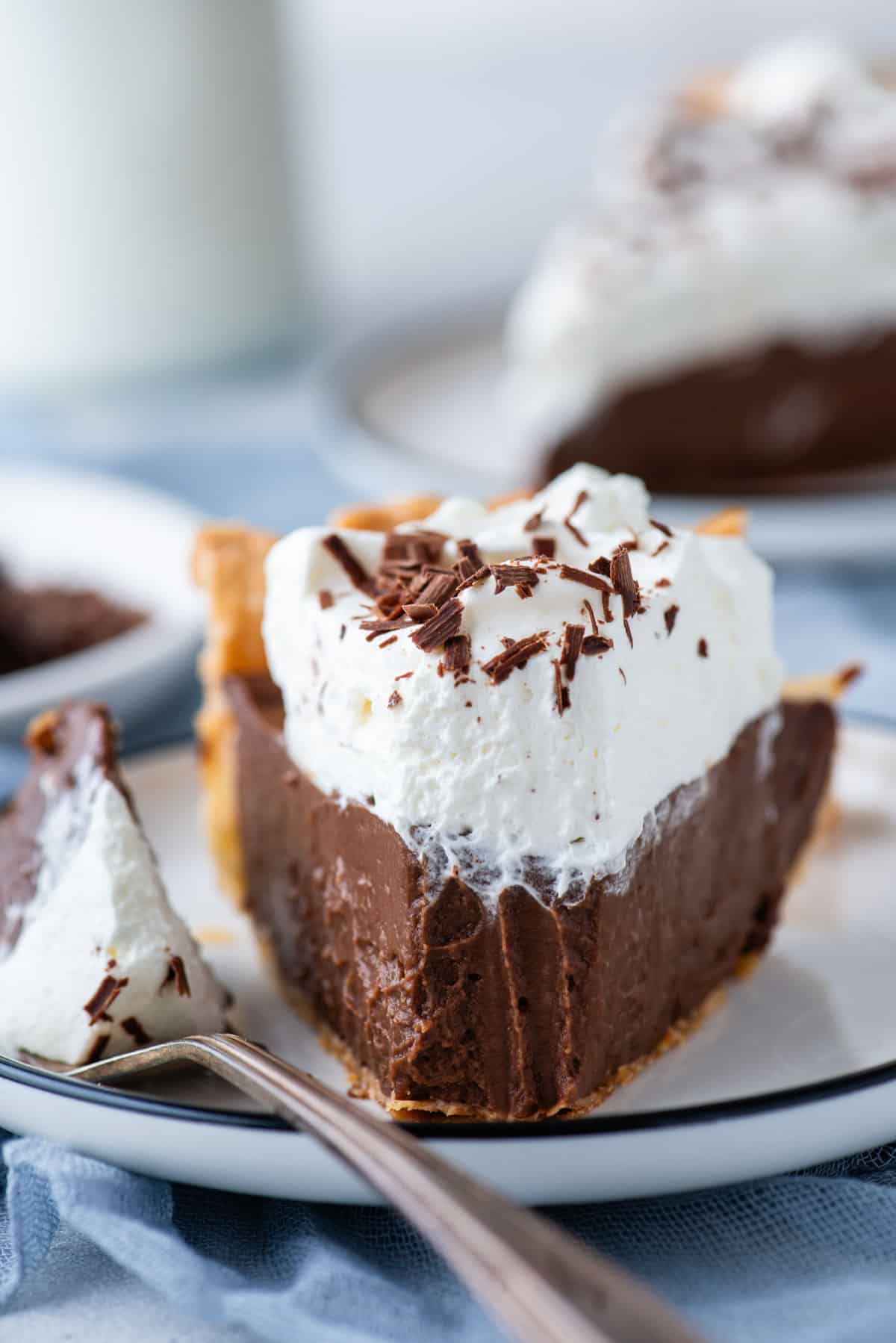 a slice of chocolate pie with a bite taken out which is laying on a fork beside the pie slice on a white plate, sitting on top of a blue towel with more pie slices in the background