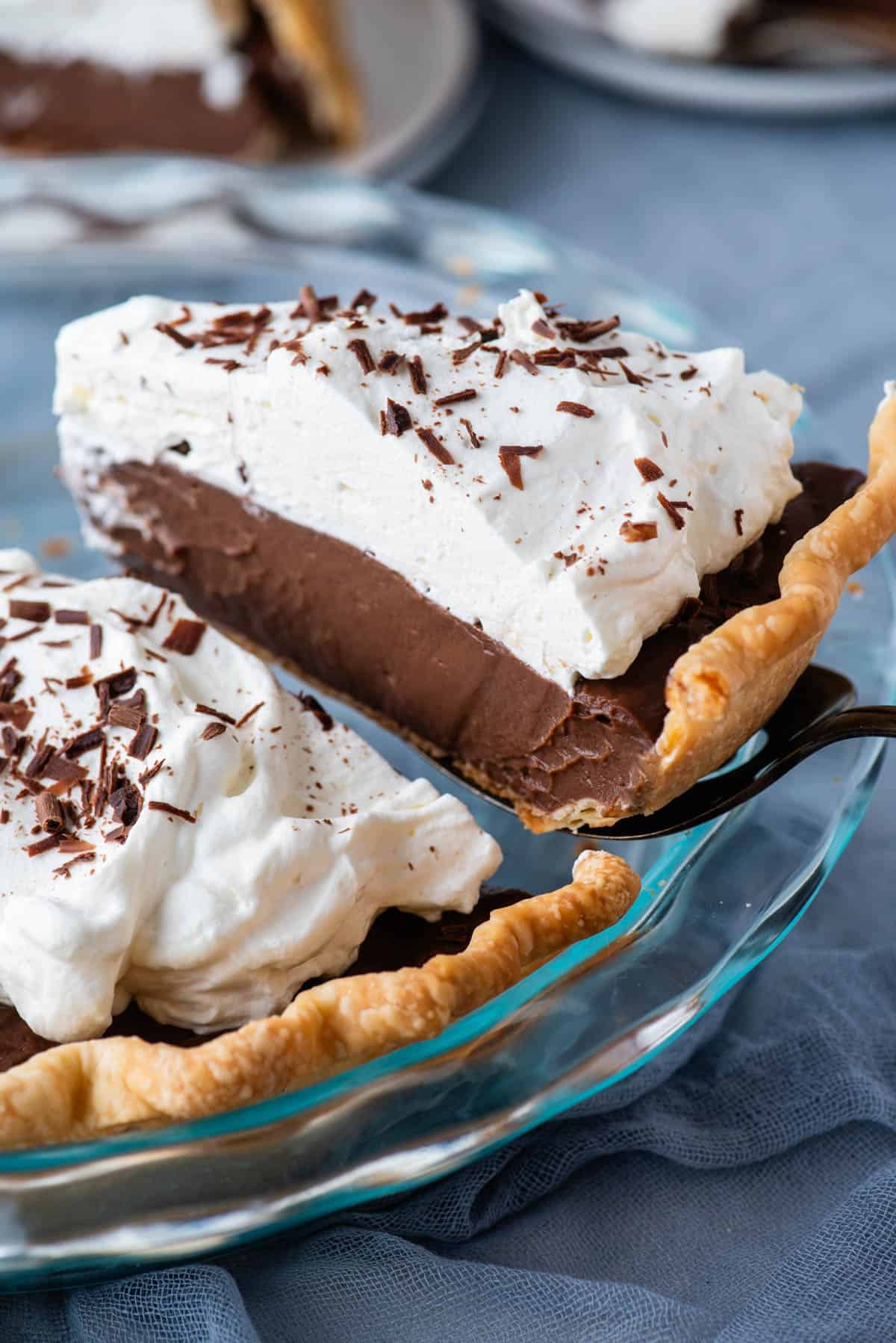a slice of chocolate pie being lifted out of a glass pie dish sitting on top of a blue towel with more pie slices on plates in the background
