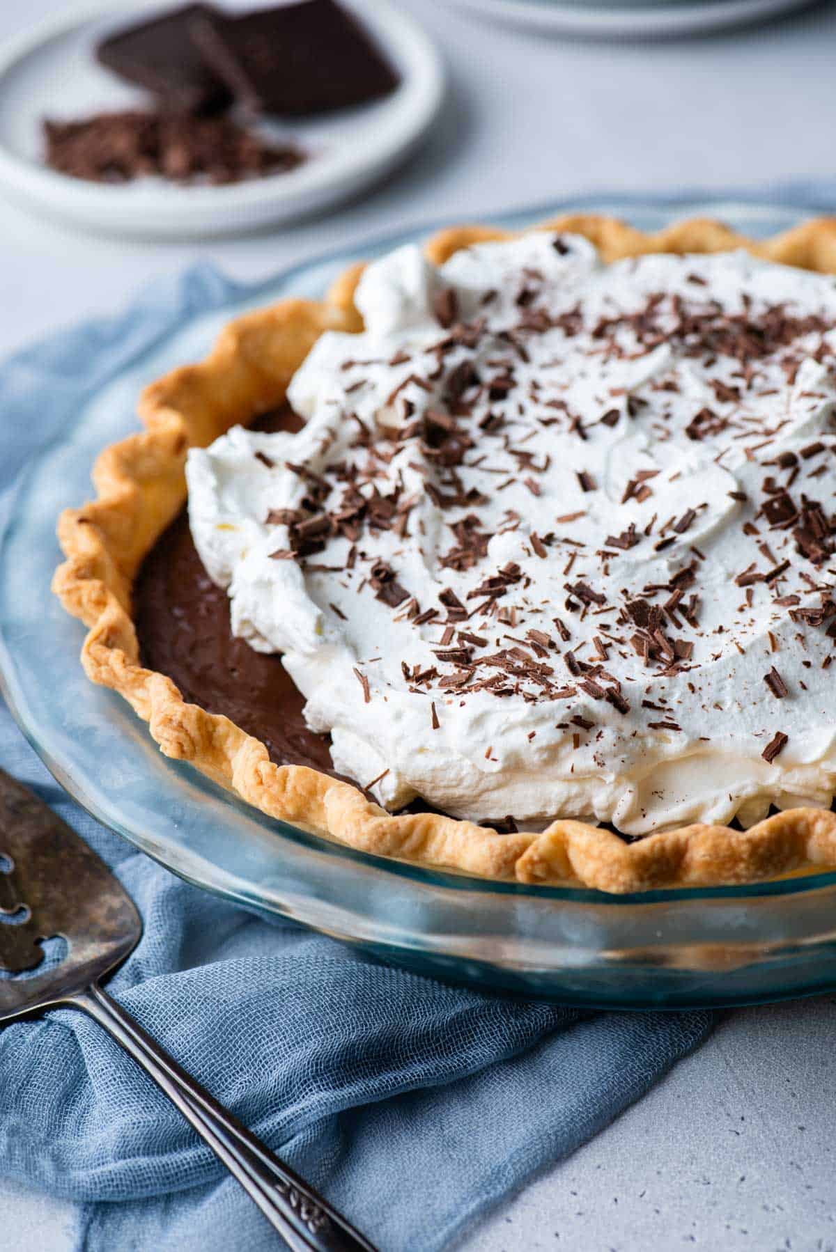 a chocolate pie in a glass pie dish sitting on a blue towel with a serving spatula beside it and a plate of chocolate in the background