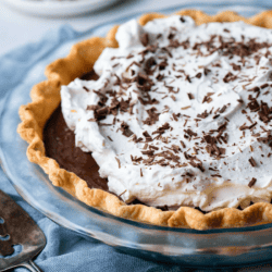 a chocolate pie topped with whipped cream and chocolate shavings in a glass pie dish on a blue towel with a serving spatula beside it