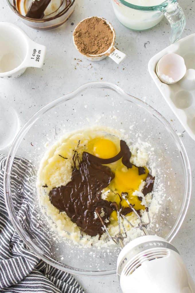 ingredients for german chocolate cake being mixed in a glass bowl with an electric mixer, surrounded by measuring cups with cocoa powder, an egg, chocolate and milk