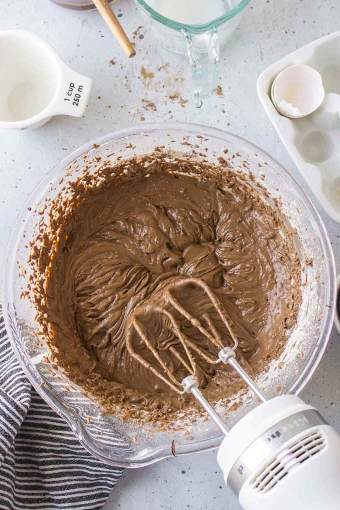 ingredients for german chocolate cake being mixed in a glass bowl with an electric mixer surrounded by a black and white striped towel, a white measuring cup, a glass measuring cup full of milk and an empty egg sheel in a white egg carton