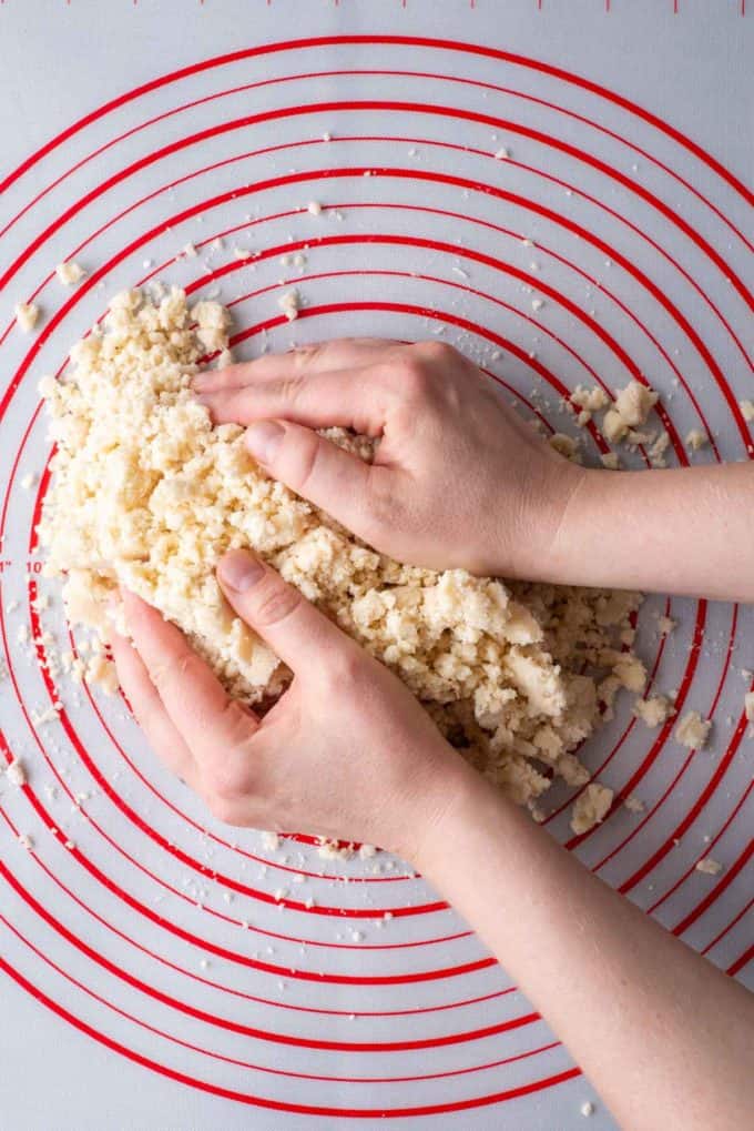 Pie crust dough that is crumbly in chunks on a red and white pie mat with two hands grabbing it, starting to form the dough