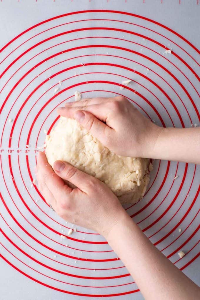 two hands forming a large disc of pie crust dough on a red and white pie mat