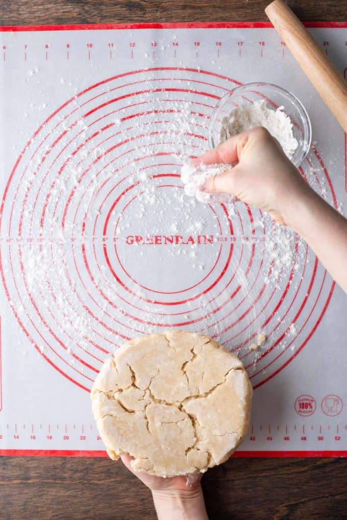 a disc of pie dough sitting on the bottom of a red and white pie mat that is being sprinkled with flour from a small glass bowl of flour and a wooden rolling pin sitting on the right side of the mat
