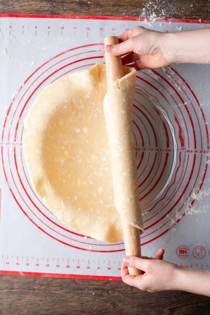 pie crust being rolled off of a rolling pin and onto a glass pie dish that is sitting on top of a red and white pie mat