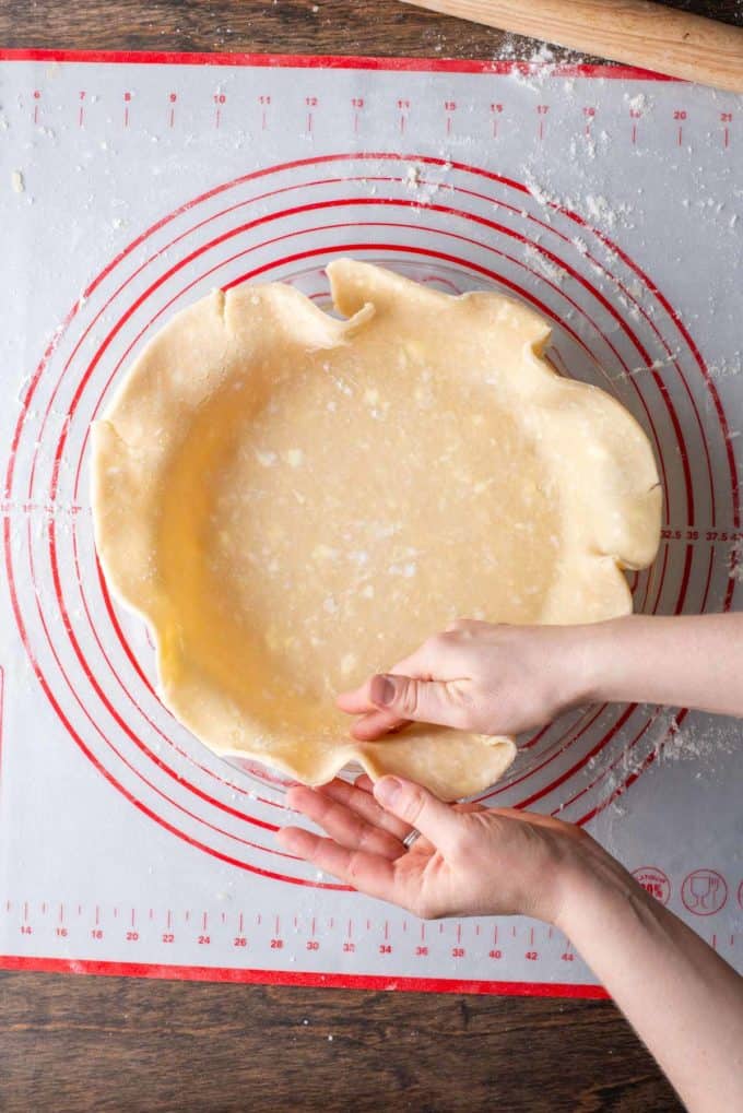 pie crust being formed by hand into a glass pie dish sitting on top of a red and white pie mat