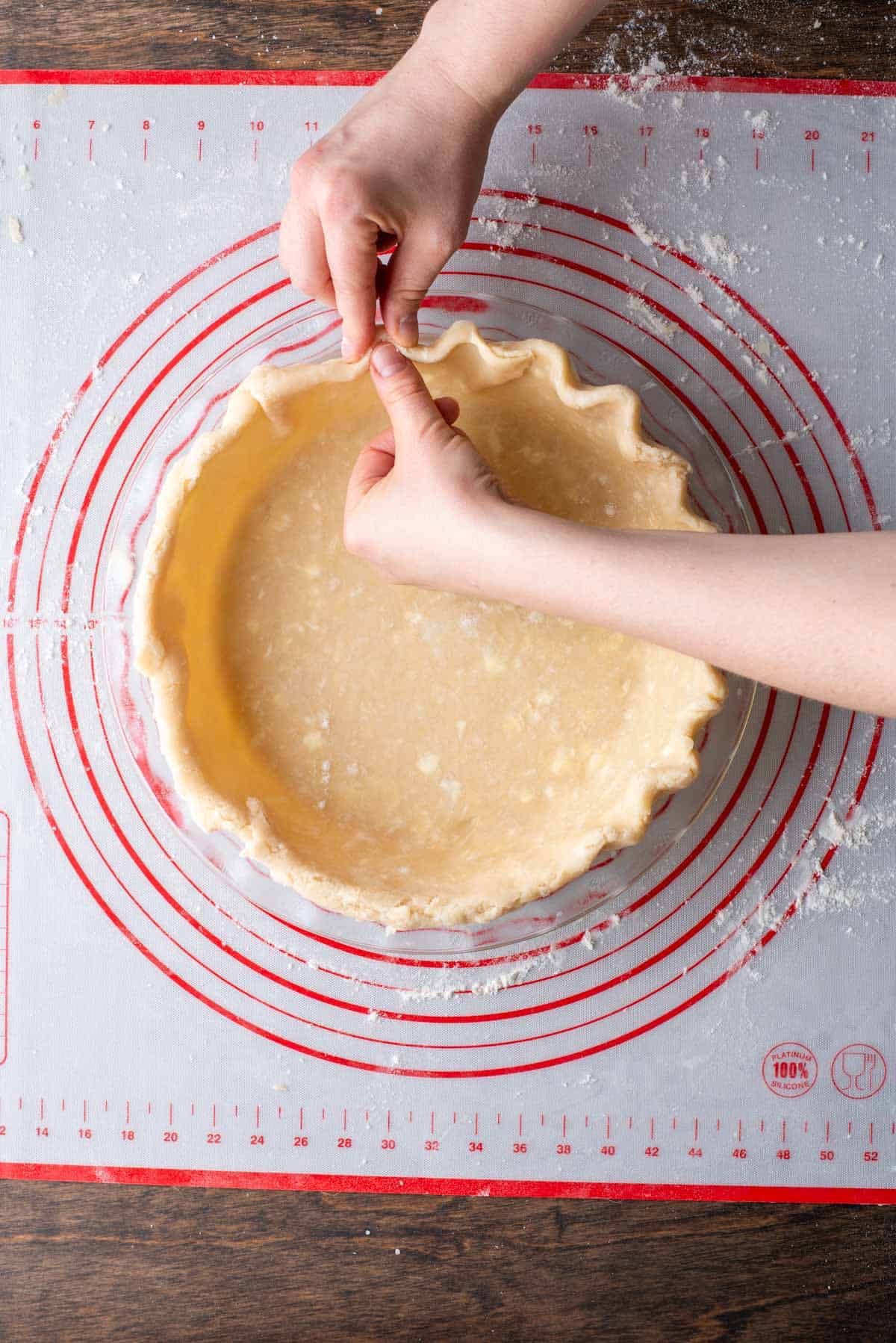 pie crust being crimped around the edges of a glass pie dish sitting on top of a red and white pie mat dusted with flour