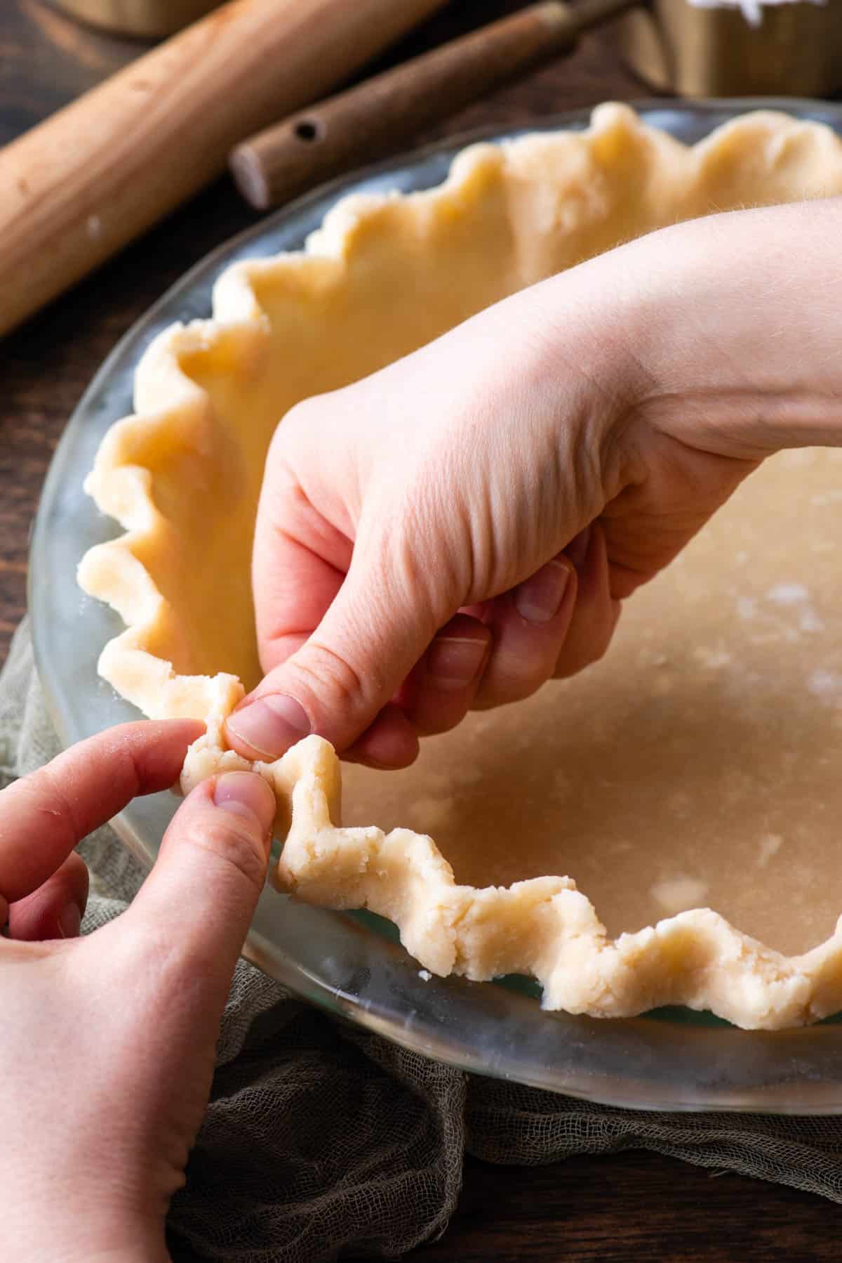 two hands crimping the edge of a pie crust in a clear glass pie pan