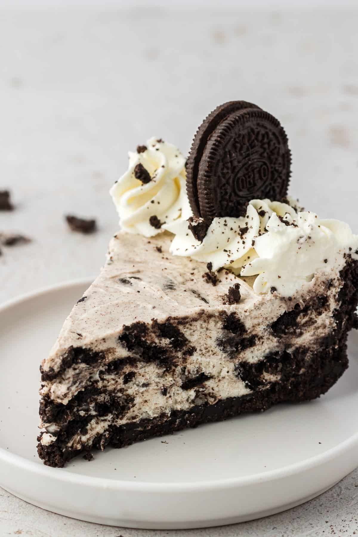 a slice of oreo pie on a white plate topped with whipped cream and a whole oreo with oreo crumbs in the back behind the plate