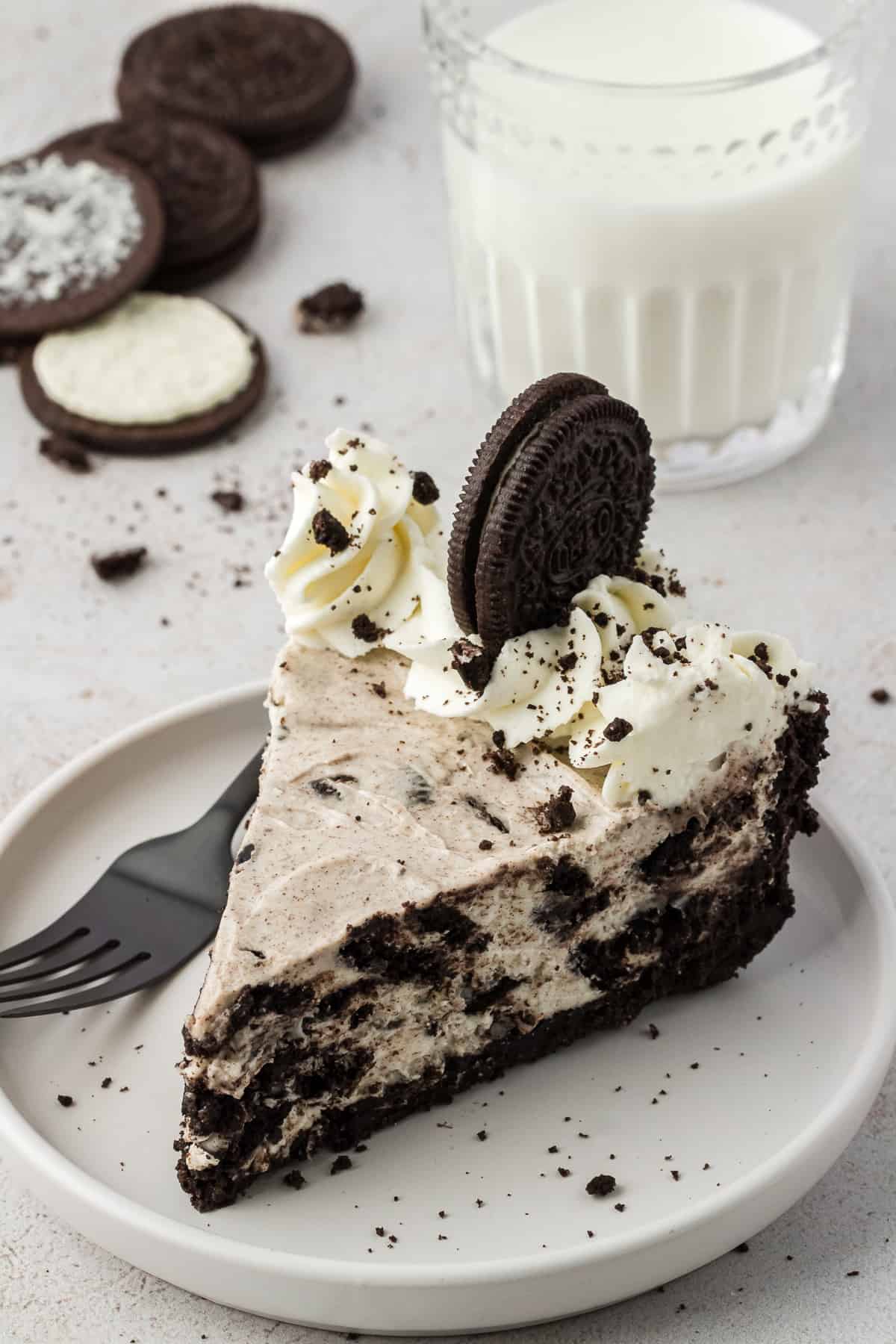 a slice of oreo pie on a white plate with a fork beside it, with a glass of milk and oreo pieces behind the plate