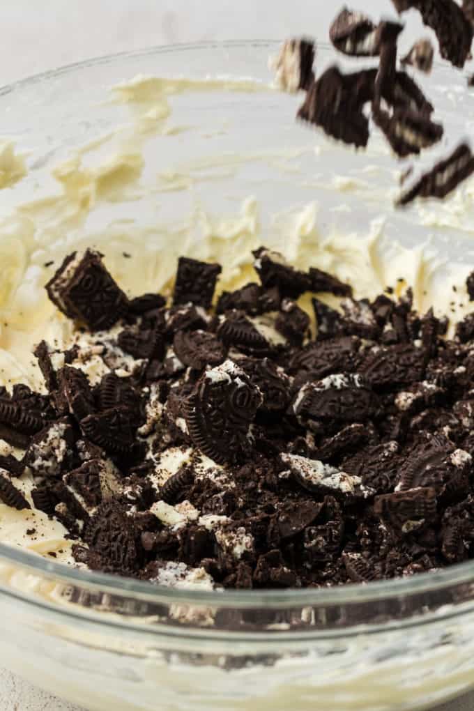 oreo pie filling in a clear glass bowl with oreo pieces being poured in on top of it