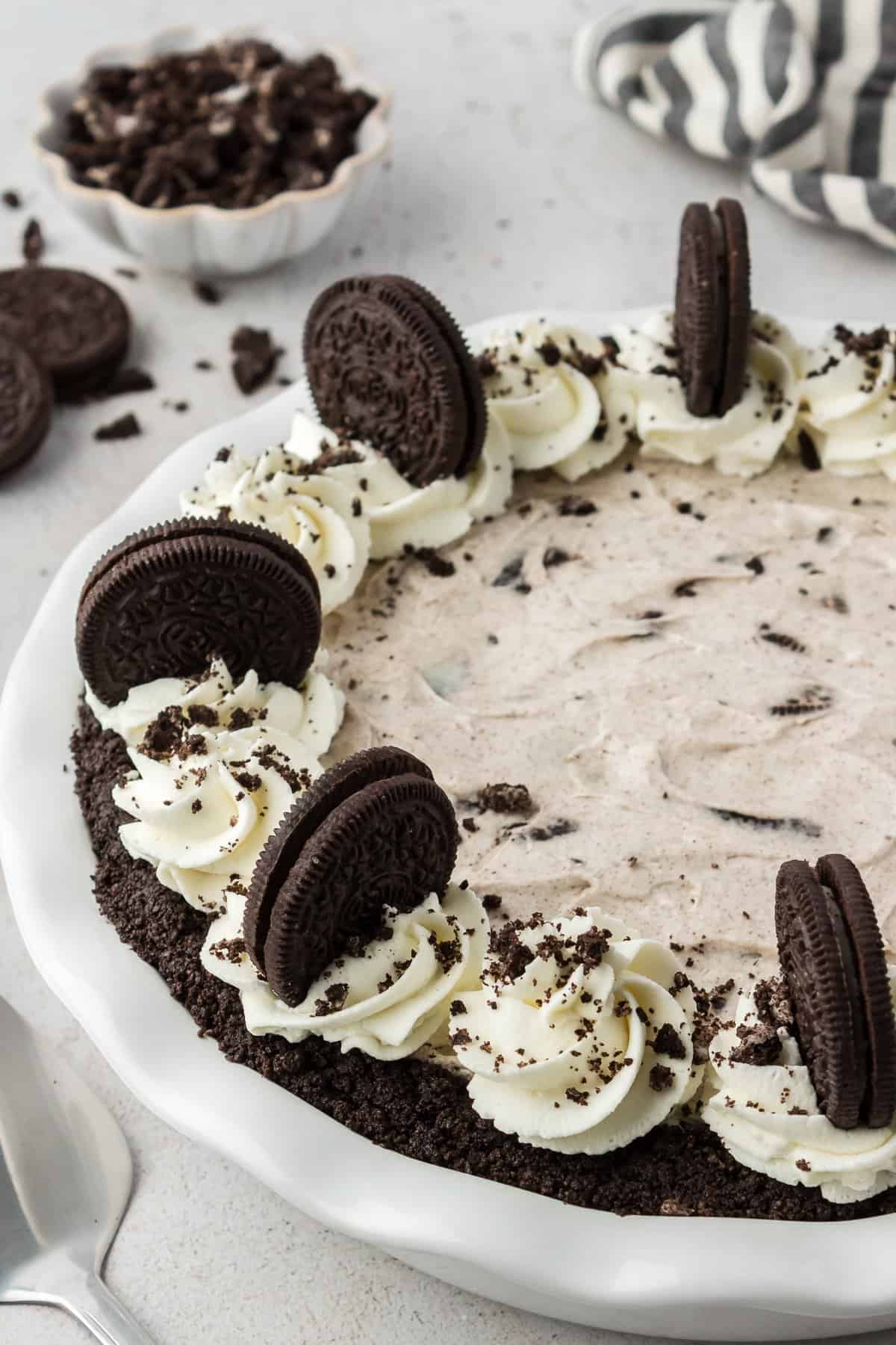 an oreo pie decorated with whipped cream and oreos on top in a white pie dish with more whole oreos and oreo pieces and a black and white striped towel in the background