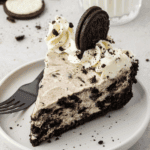 A slice of no bake oreo pie cheesecake on a white plate, topped with whipped cream, cookie crumbles, and a whole chocolate sandwich cookie, rests beside a black fork. A glass of milk is in the background.