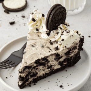 A slice of no bake oreo pie cheesecake on a white plate, topped with whipped cream, cookie crumbles, and a whole chocolate sandwich cookie, rests beside a black fork. A glass of milk is in the background.