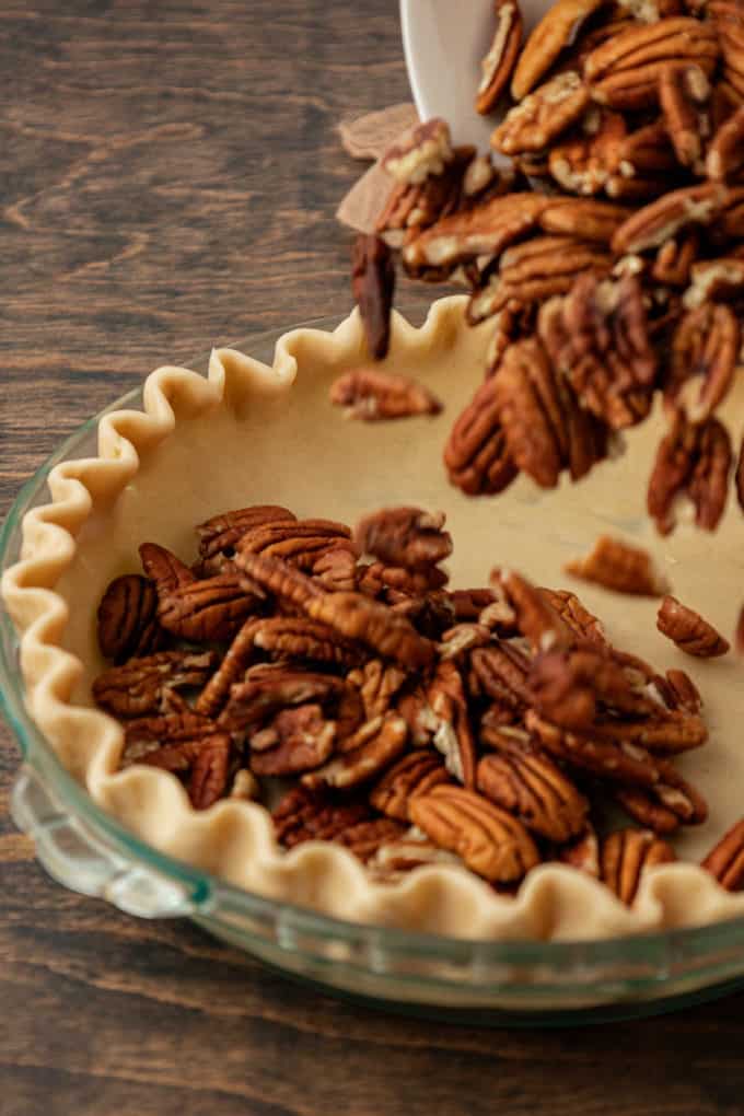 pecans being added to a glass pie dish with unbaked pie crust in it sitting on a wood surface